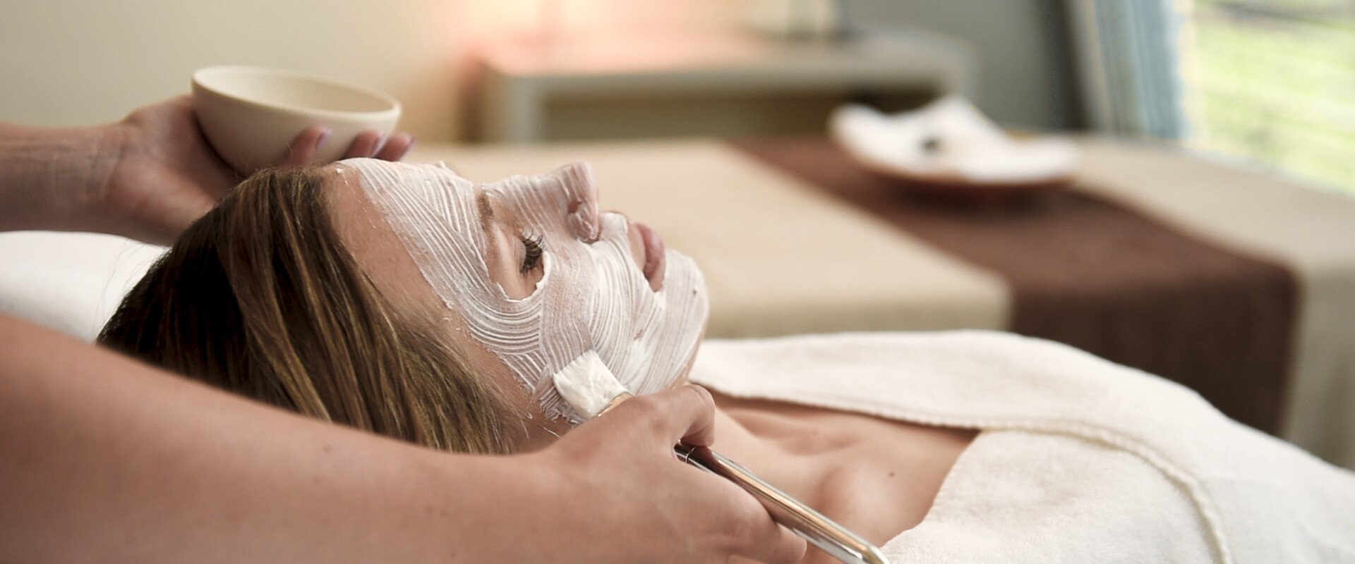 Eine Person, die auf einer Wellnessliege liegt, bekommt in entspannter Atmosphäre von einem Therapeuten eine weiße Gesichtsmaske mit einem Pinsel aufgetragen.