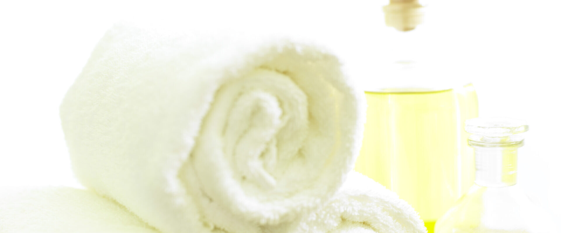 Drei gerollte weiße Handtücher mit zwei Glasflaschen mit gelbem Öl in einer hellen, Spa-ähnlichen Umgebung.