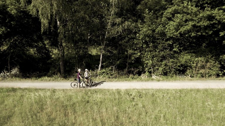 Zwei Personen mit Fahrrädern gehen an einem sonnigen Tag einen Feldweg entlang, der an dichten grünen Bäumen und Gras vorbeiführt.