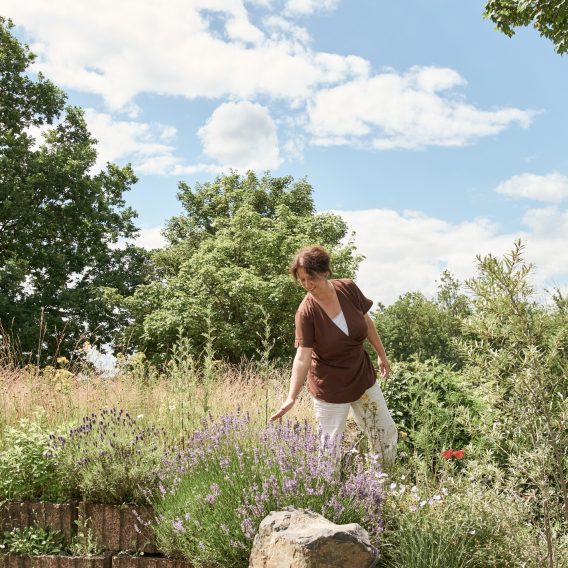 Eine Person pflegt einen üppigen Garten mit violetten Blumen und Grün unter einem sonnigen, teilweise bewölkten Himmel.