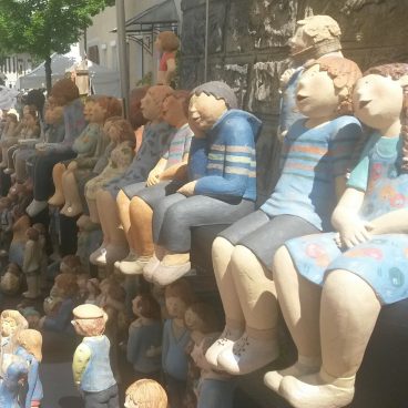 Reihen bunter Keramikfiguren von Menschen, die dicht beieinander sitzen und an einem sonnigen Tag im Freien ausgestellt sind.