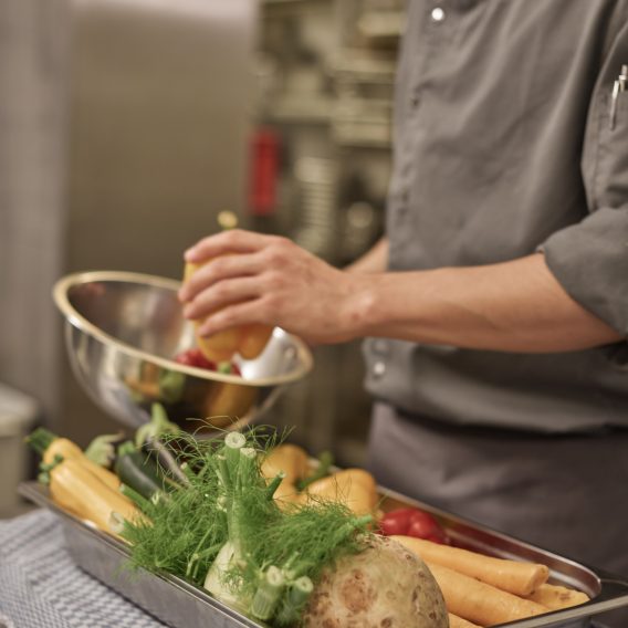 Koch in grauer Uniform bereitet Gemüse zu, mit Wurzelgemüse und Fenchel auf einem Tablett auf einer Küchentheke.