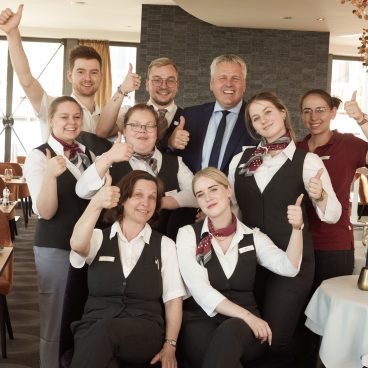 Eine Gruppe lächelnder Restaurantmitarbeiter in Uniform streckt in einem Essbereich gemeinsam den Daumen nach oben.