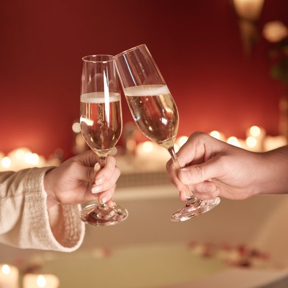 Zwei Personen stoßen vor einem Bad bei Kerzenschein mit Champagner an und schaffen so eine romantische und gemütliche Atmosphäre.