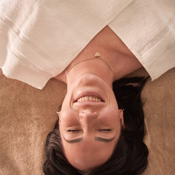 Eine Frau liegt auf einem Massagetisch, ist mit einem weißen Handtuch bedeckt, lächelt mit geschlossenen Augen und wirkt entspannt und zufrieden.