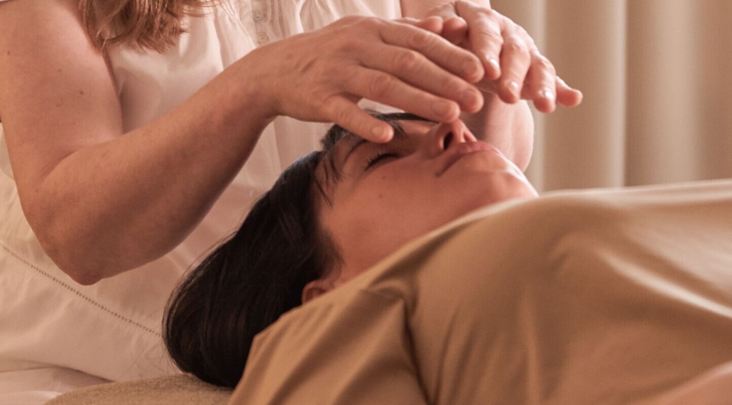 Eine Frau führt Energieheilung oder Reiki durch, indem sie ihre Hände über dem Gesicht einer anderen Frau schweben lässt, während diese auf einem Behandlungstisch liegt.