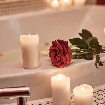 Eine rote Rose und brennende weiße Kerzen werden auf den Rand einer Badewanne gestellt und schaffen eine romantische, gemütliche Atmosphäre.