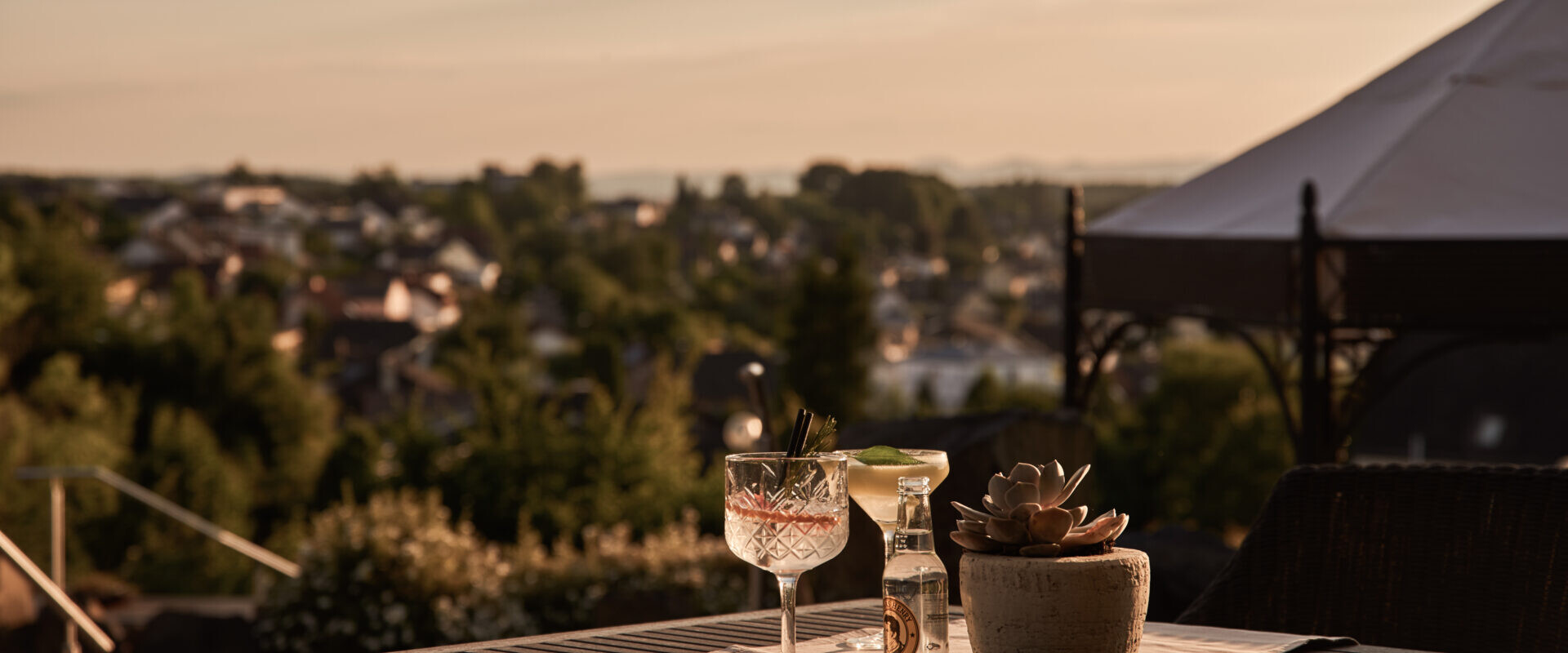 Ein Tisch mit Cocktails und einer Topfpflanze bietet Aussicht auf eine malerische Stadt bei Sonnenuntergang, mit sanftem goldenen Licht und einem Pavillon im Hintergrund.