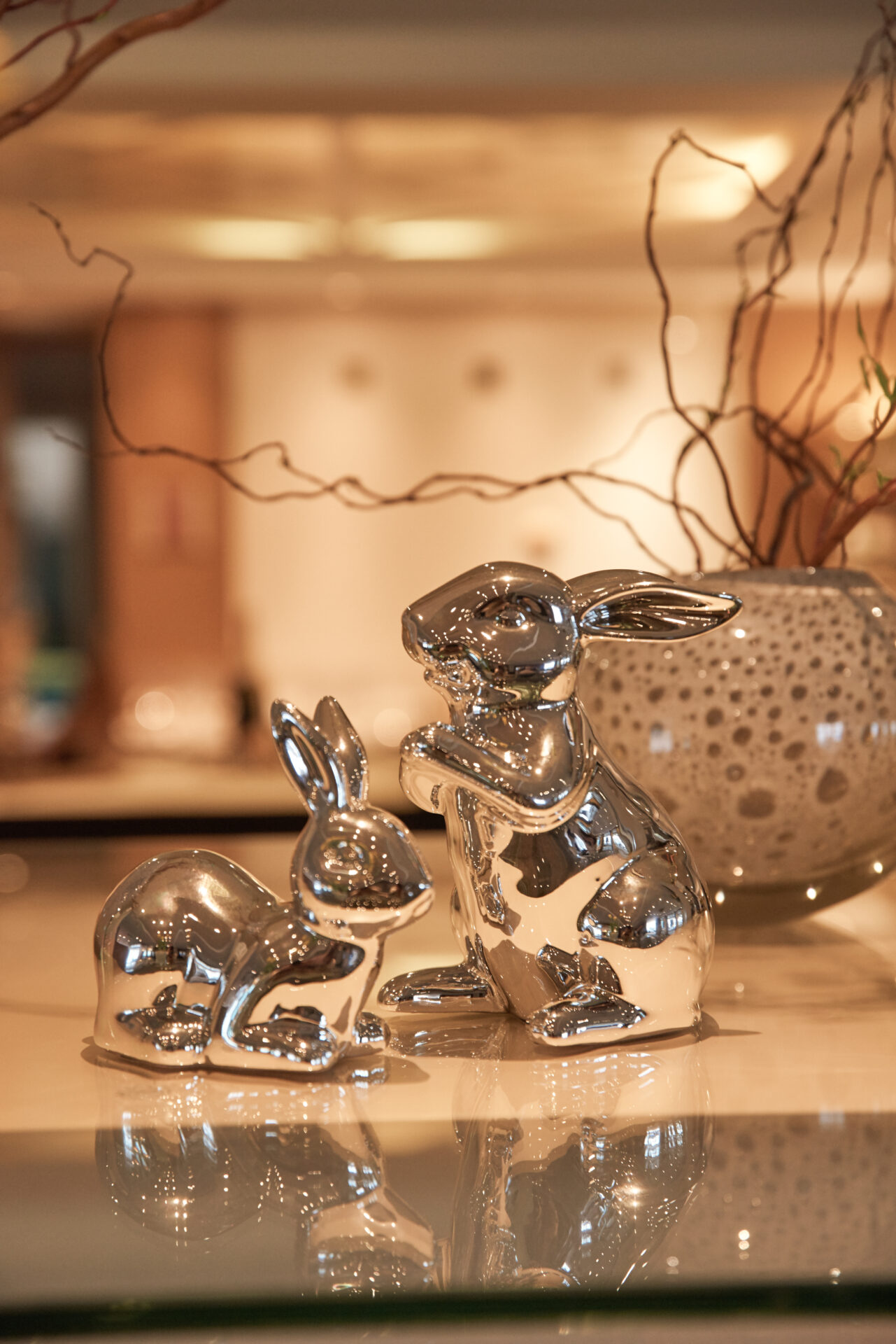 Auf einer Fläche sind zwei glänzende silberne Hasenfiguren ausgestellt, im Hintergrund eine dekorative Vase und Zweige.