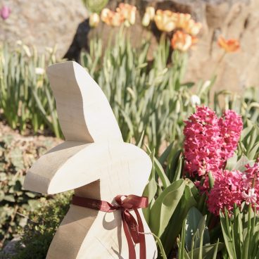 Ein Holzhase mit einer roten Schleife sitzt in einem Garten zwischen rosa Hyazinthen und gelben Tulpen, im Hintergrund sind Felsen.