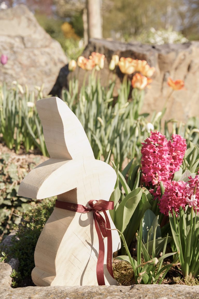 Ein Holzhase mit einer roten Schleife sitzt in einem Garten zwischen rosa Hyazinthen und gelben Tulpen, im Hintergrund sind Felsen.