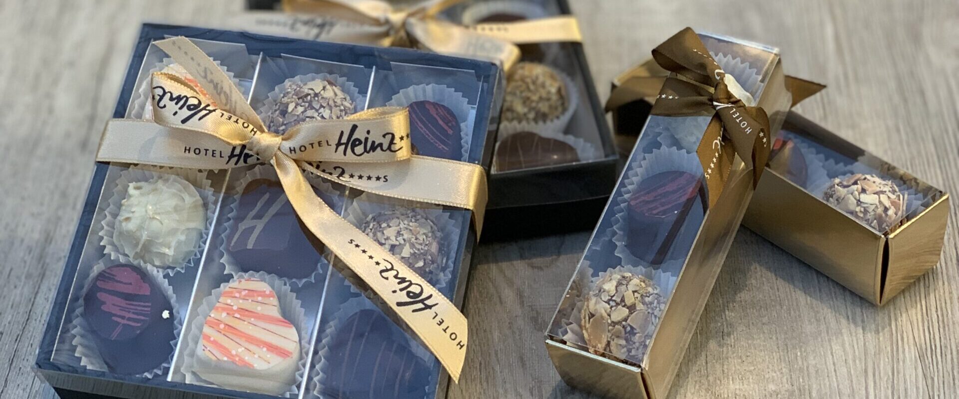 Zwei Schachteln mit verschiedenen Pralinen und zwei kleinere Schachteln, alle mit goldenen Bändern zusammengebunden, auf einer Holzoberfläche präsentiert.