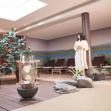 Eine Frau im Bademantel steht in einer modernen Spa-Lounge mit Topfpflanzen, Liegestühlen und sanftem Tageslicht.