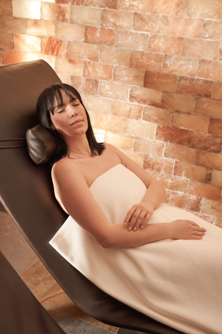 Eine in ein weißes Handtuch gehüllte Frau entspannt auf einem Liegestuhl in einem Spa-Raum mit einer Salzziegelwand.