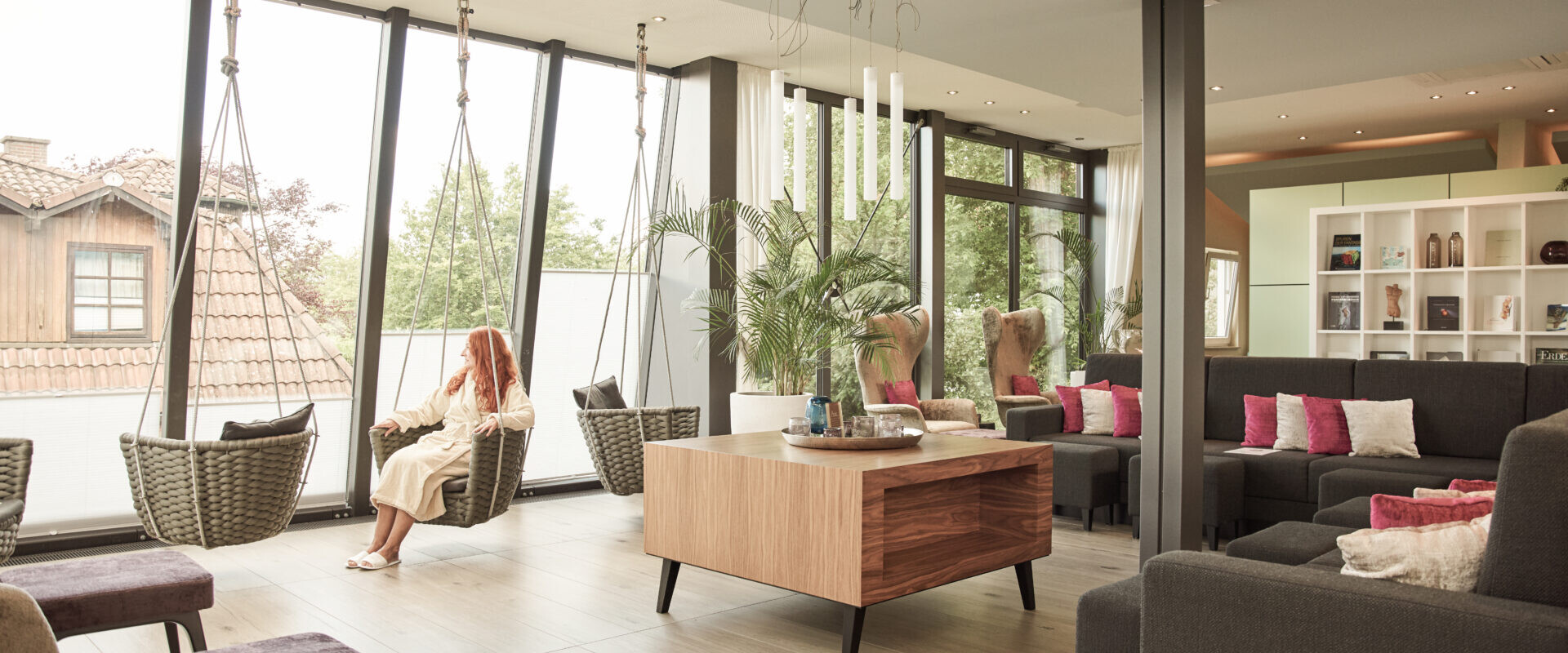 Frau im Bademantel sitzt auf einem Hängesessel in einer hellen, modernen Lounge mit großen Fenstern und gemütlichen Sitzgelegenheiten.