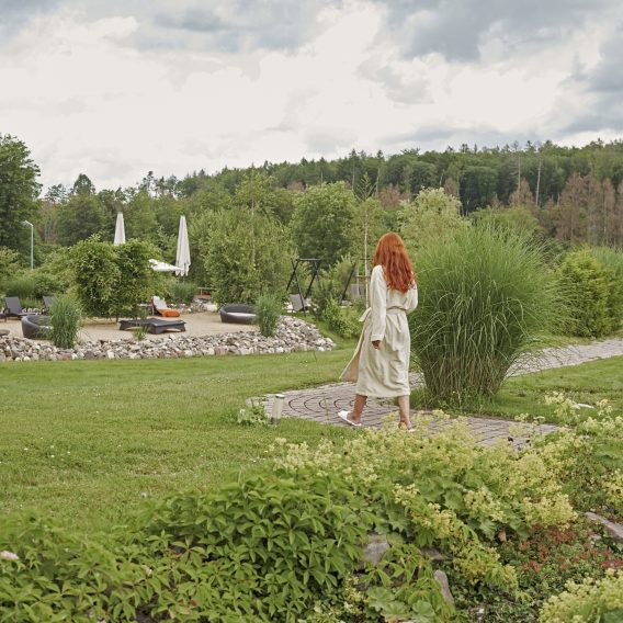 Eine Frau mit langen roten Haaren geht auf einem Gartenweg auf einen Loungebereich im Freien zu, umgeben von üppigem Grün und bewölktem Himmel.