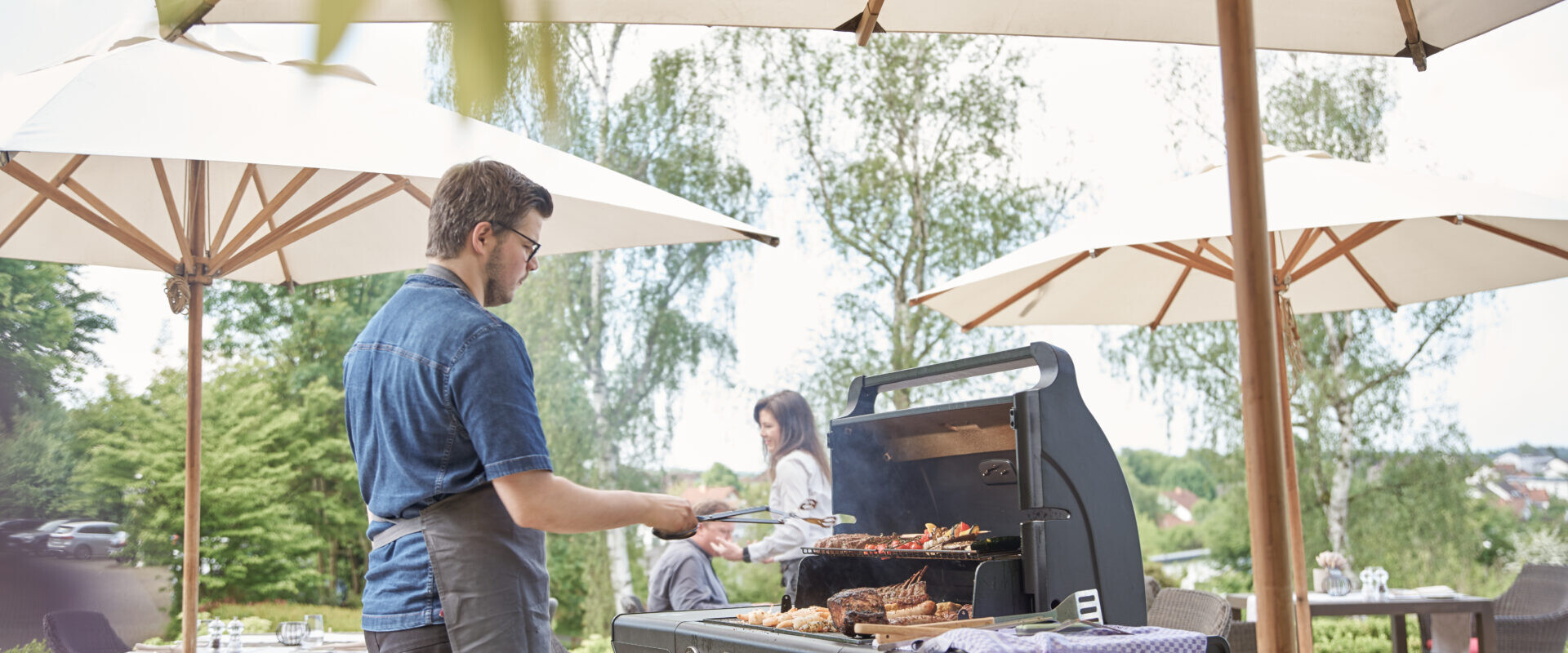 An einem sonnigen Tag grillt ein Mann im Freien unter großen Sonnenschirmen Essen, im Hintergrund stehen Tische und eine weitere Person.