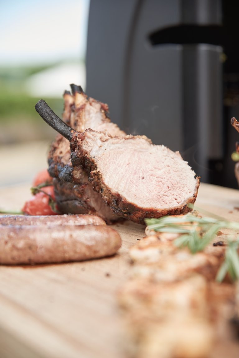 Nahaufnahme eines in Scheiben geschnittenen, gegrillten Schweinerückens mit Würstchen und Kräutern auf einem Holzbrett im Freien.