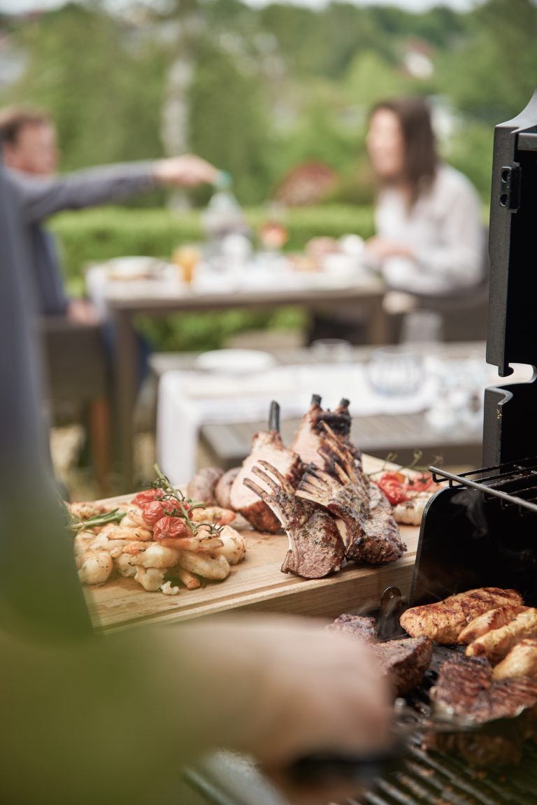 Eine Person grillt Fleisch im Freien, mit gekochtem Essen auf einem Holzbrett und Menschen, die im unscharfen Hintergrund speisen.