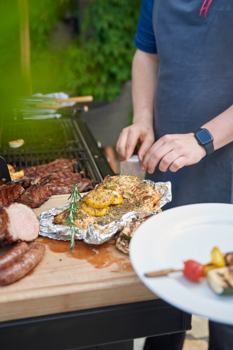 Person grillt Fleisch und Gemüse im Freien und bereitet das Essen auf einem Holzbrett neben einem Grill zu.