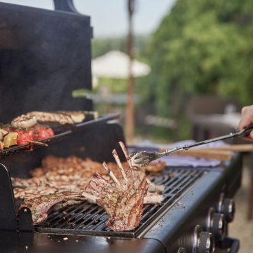 Eine Person grillt Fleisch, Gemüse und ein Lammkarree auf einem Grill im Freien, mit einem großen Sonnenschirm und Grün im Hintergrund.