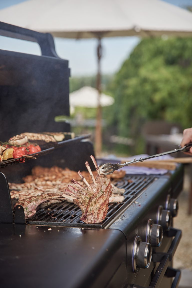 Eine Person grillt Fleisch, Gemüse und ein Lammkarree auf einem Grill im Freien, mit einem großen Sonnenschirm und Grün im Hintergrund.