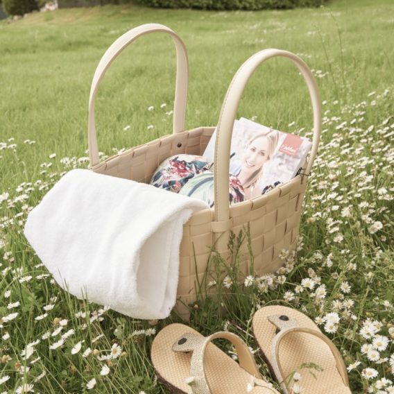 Ein geflochtener Picknickkorb mit einem weißen Handtuch, einer Zeitschrift und zwei Paar Sandalen steht auf Gras und Gänseblümchen.