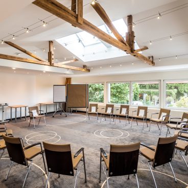 Ein geräumiger, gut beleuchteter Tagungsraum mit kreisförmig angeordneten Stühlen, großen Fenstern und freiliegenden Holzbalken an der Decke.