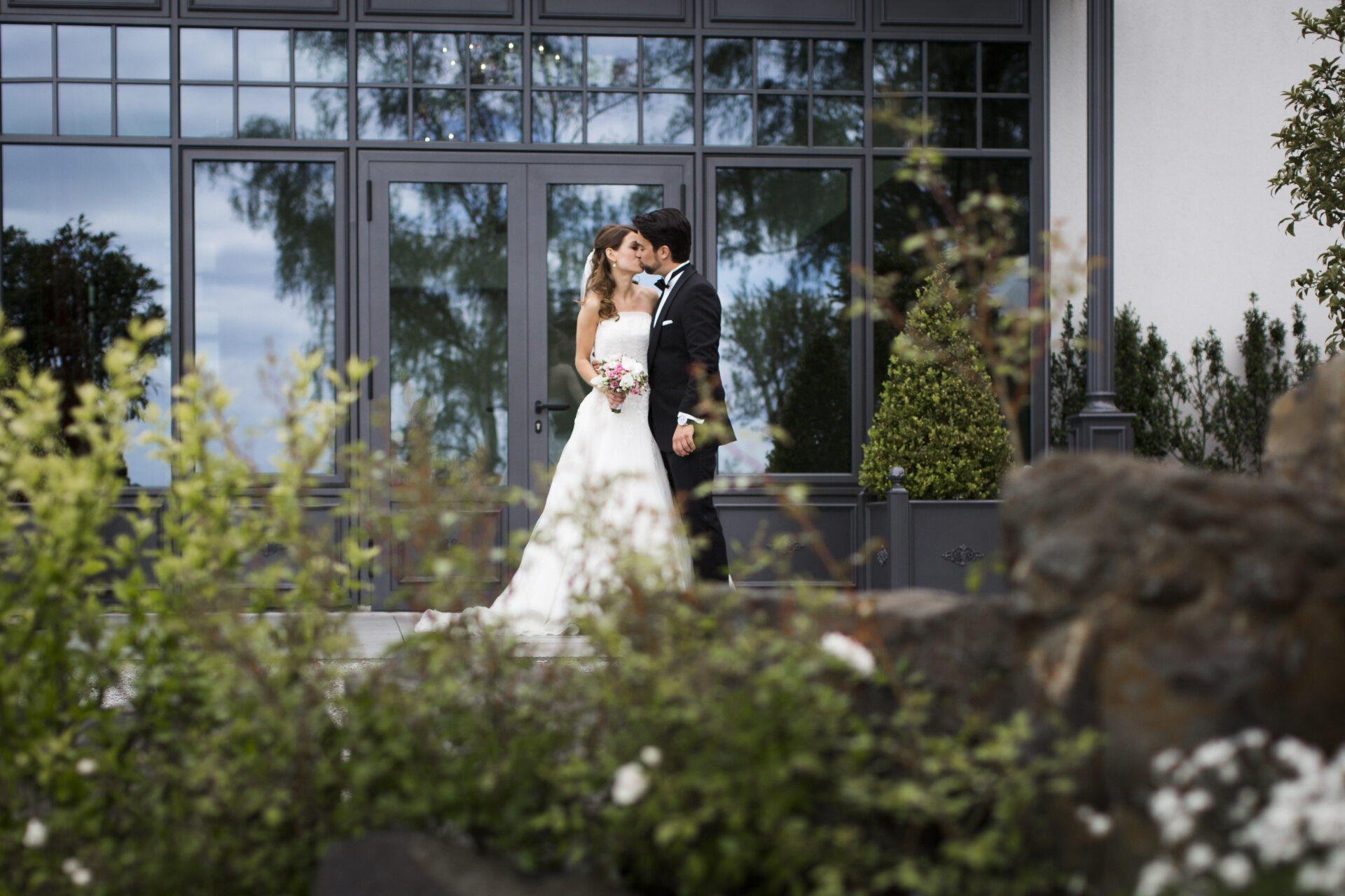 An ihrem Hochzeitstag küssen sich Braut und Bräutigam draußen vor großen Fenstern, umgeben von Grün und Blumen.