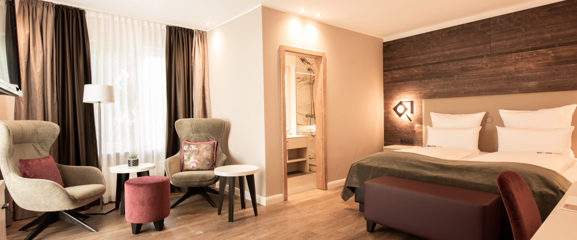 Modernes Hotelzimmer mit Doppelbett, Sesseln, kleinem Tisch, Holzboden und offenem Badezimmerbereich.