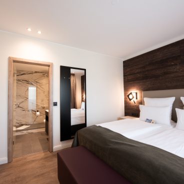Modernes Hotelzimmer mit großem Bett, Akzentwand aus dunklem Holz und Blick durch eine offene Tür in ein Badezimmer mit Marmorfliesen.