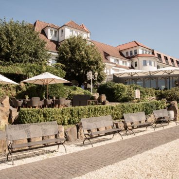 Eine sonnige Außenterrasse mit Bänken, Tischen und Sonnenschirmen vor einem großen Gebäude mit rotem Dach und weißen Wänden.