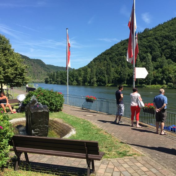 An einem sonnigen Tag stehen und sitzen Menschen in einem Park am Flussufer mit Fahnen, üppig grünen Hügeln und einem Brunnen.