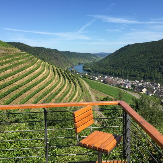 Terrassenförmige Weinberge und ein Flusstal mit einem Dorf, von einem Balkon mit einem Holzstuhl unter einem klaren blauen Himmel betrachtet.
