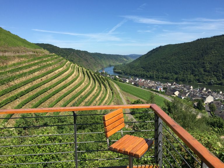 Von einem Holzstuhl auf einem Balkon aus hat man Ausblick auf terrassierte Weinberge, einen Fluss und ein zwischen grünen Hügeln eingebettetes Dorf unter einem klaren blauen Himmel.