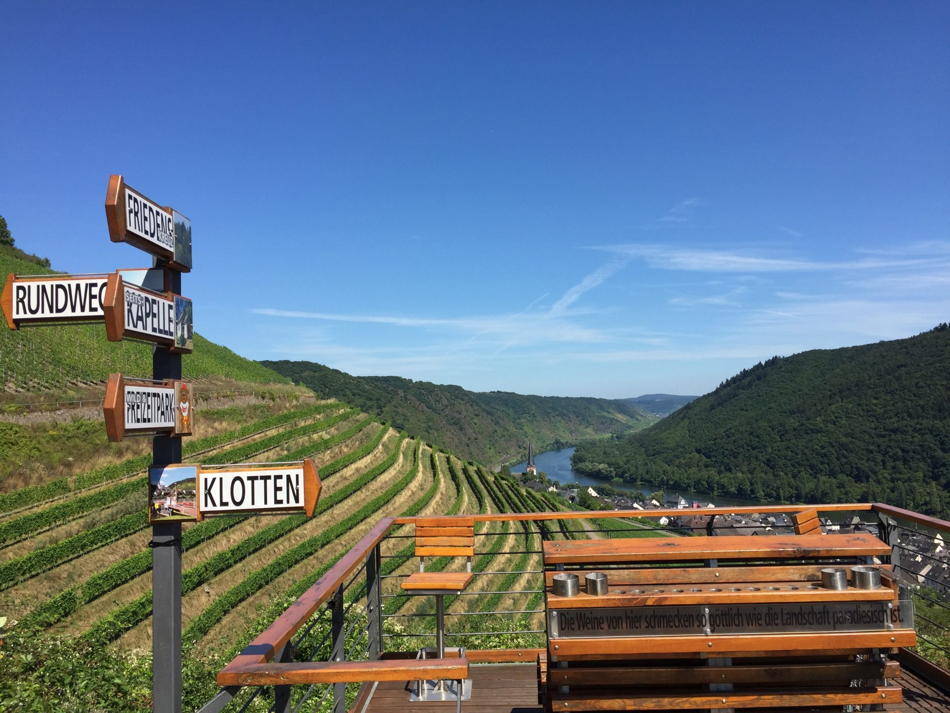 Von Holzbänken und Wegweisern aus hat man einen Blick auf grüne Weinberge auf terrassierten Hängen unter einem klaren blauen Himmel neben einem Flusstal.