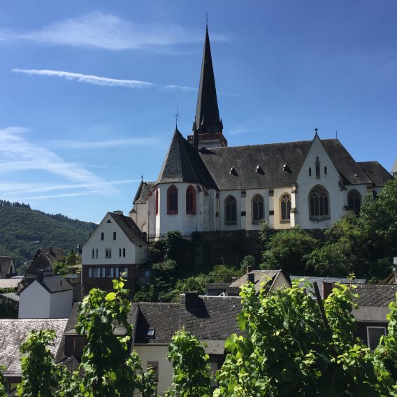 Eine große Kirche mit einem hohen Kirchturm steht zwischen Häusern und grünen Hügeln unter einem klaren blauen Himmel.