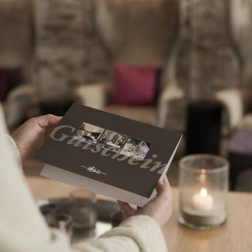 Eine Person hält eine Gästeeinladungskarte neben einer brennenden Kerze auf einem Holztisch in einer gemütlichen, sanft beleuchteten Lounge.