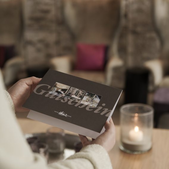 Eine Person hält eine Gästeeinladungskarte neben einer brennenden Kerze auf einem Holztisch in einer gemütlichen, sanft beleuchteten Lounge.