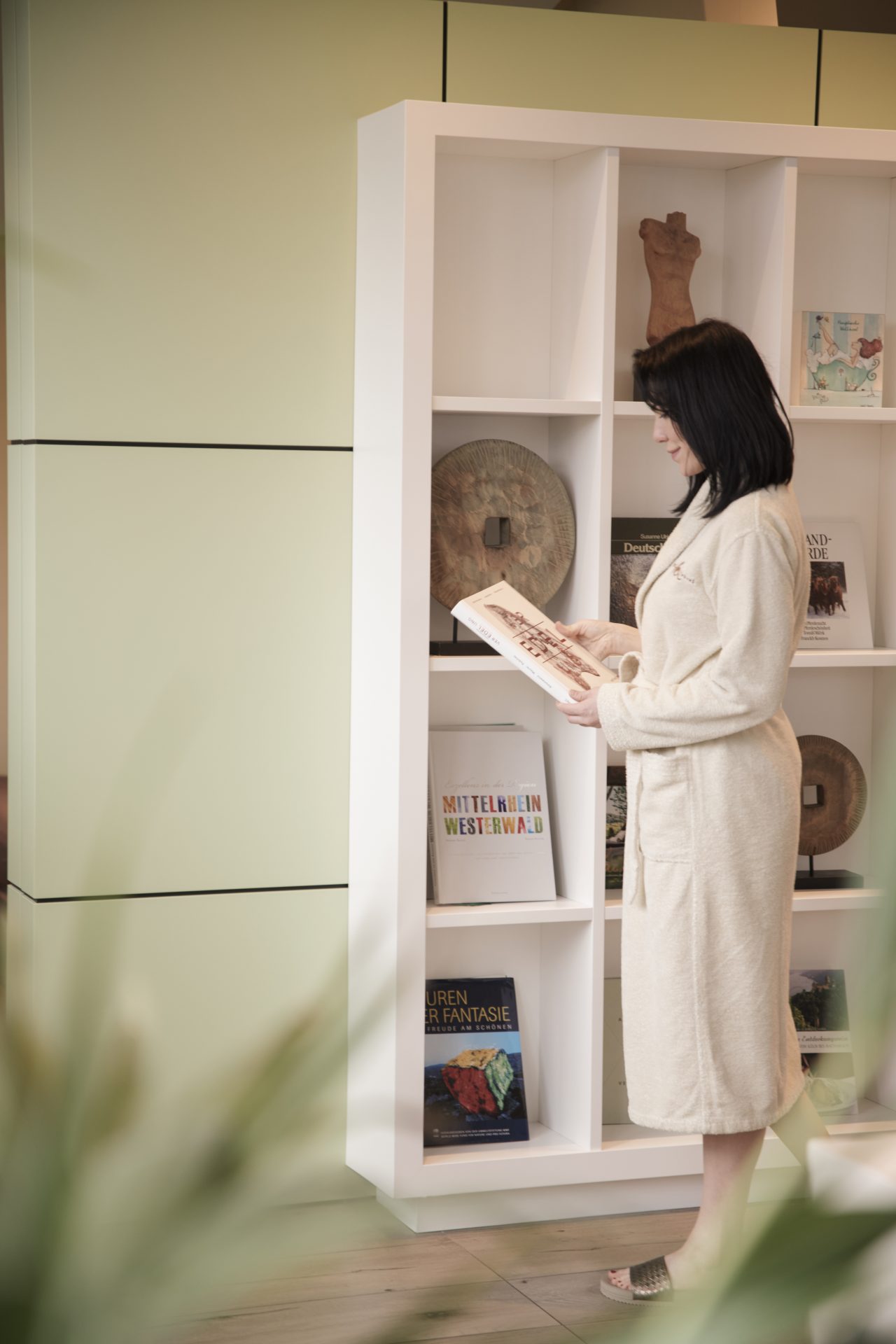 Eine Frau im Bademantel liest ein Buch, während sie neben einem modernen weißen Bücherregal voller Bücher und Dekorationsgegenstände steht.