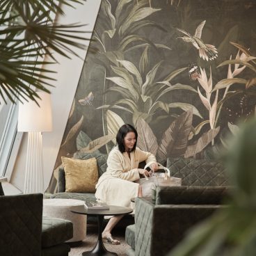 Eine Frau im Bademantel sitzt auf einem grünen Sofa in einem Wohnzimmer mit tropischer Tapete und modernem Dekor und ordnet Gegenstände in ihrer Tasche.