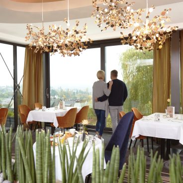 Ein Paar steht an großen Fenstern in einem modernen Restaurant und blickt nach draußen; die Tische sind gedeckt und grüne Pflanzen schmücken den Vordergrund.