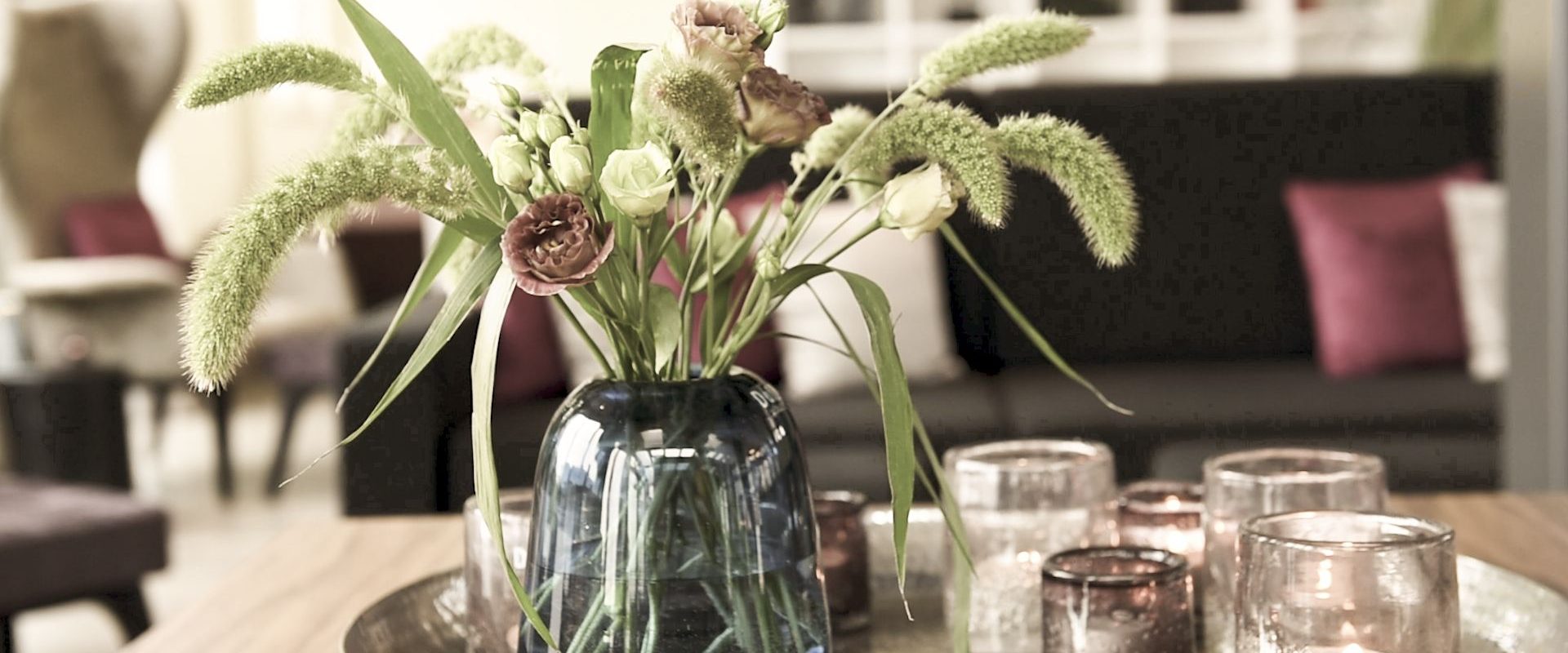 Eine blaue Glasvase mit Blumen und Gräsern steht auf einem Tablett mit Kerzenhaltern aus Glas auf einem Holztisch in einem gemütlichen Wohnzimmer.