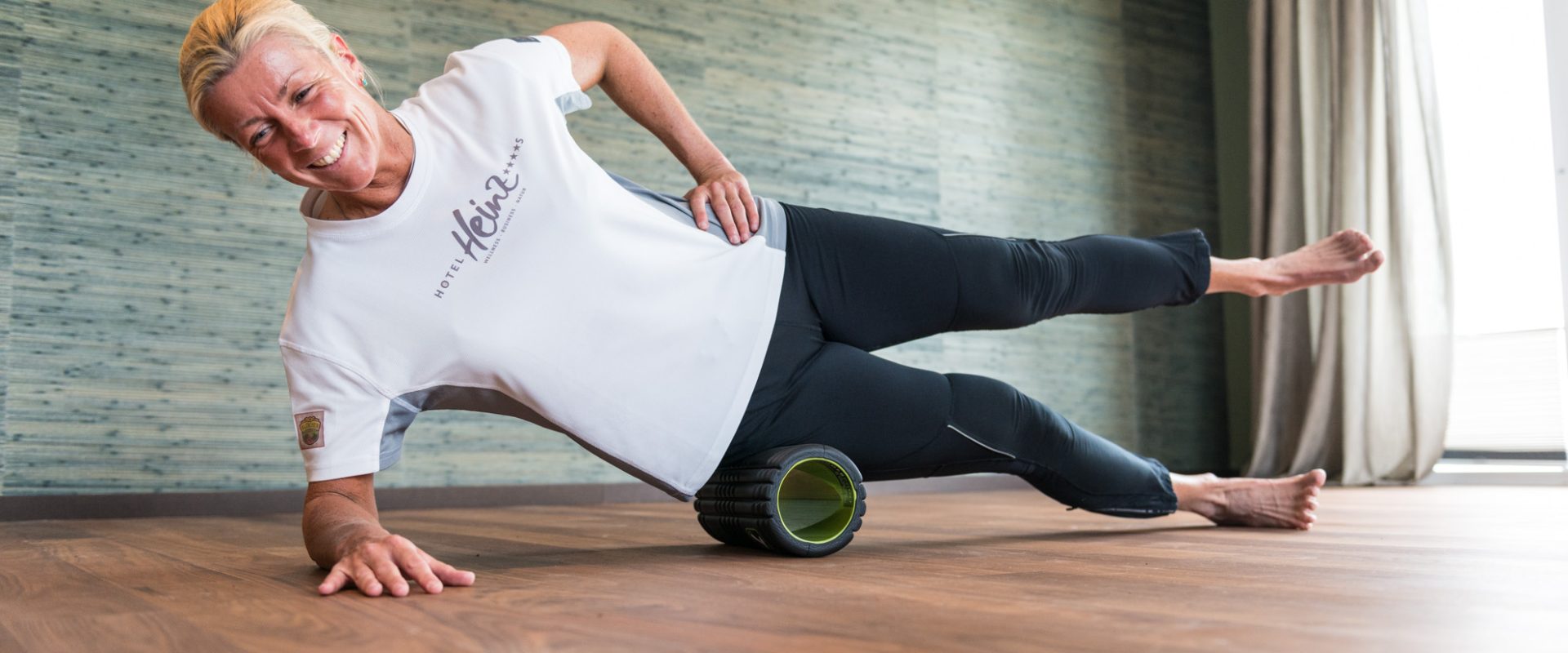 Lächelnde Frau macht eine seitliche Planke auf einer Schaumstoffrolle in einem hellen Raum mit Holzboden und grünen Wänden.