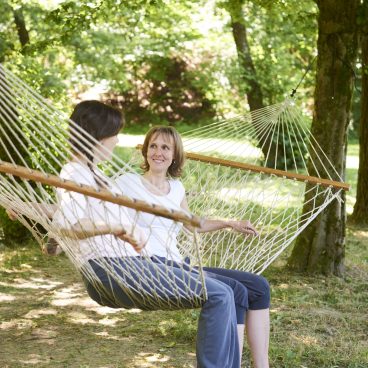 Zwei Frauen sitzen zusammen auf einer Seilhängematte im Freien, lächeln und unterhalten sich im Schatten grüner Bäume.