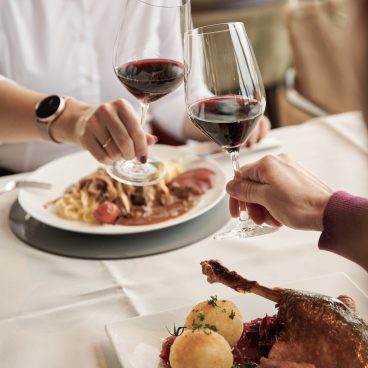 Zwei Personen stoßen mit Rotweingläsern an einem Tisch an, auf dem Teller mit Gourmetspeisen stehen, darunter Pasta und ein Fleischgericht.