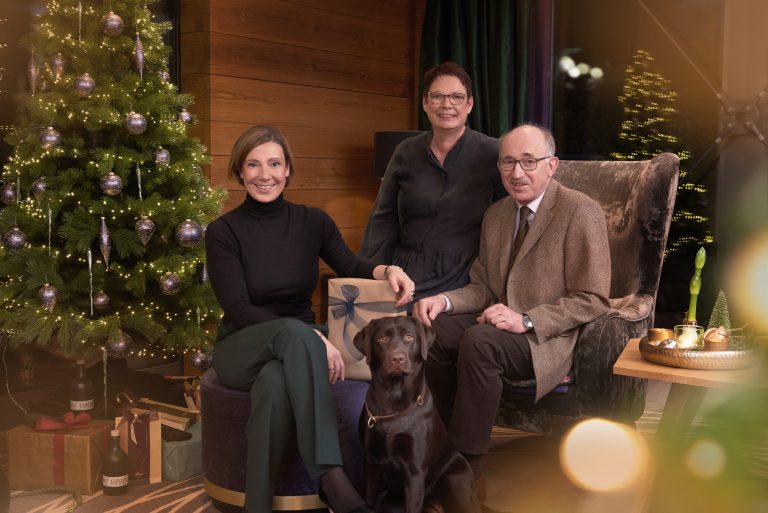 Drei Erwachsene und ein schwarzer Labrador sitzen mit Geschenken an einem geschmückten Weihnachtsbaum und lächeln herzlich in die Kamera.