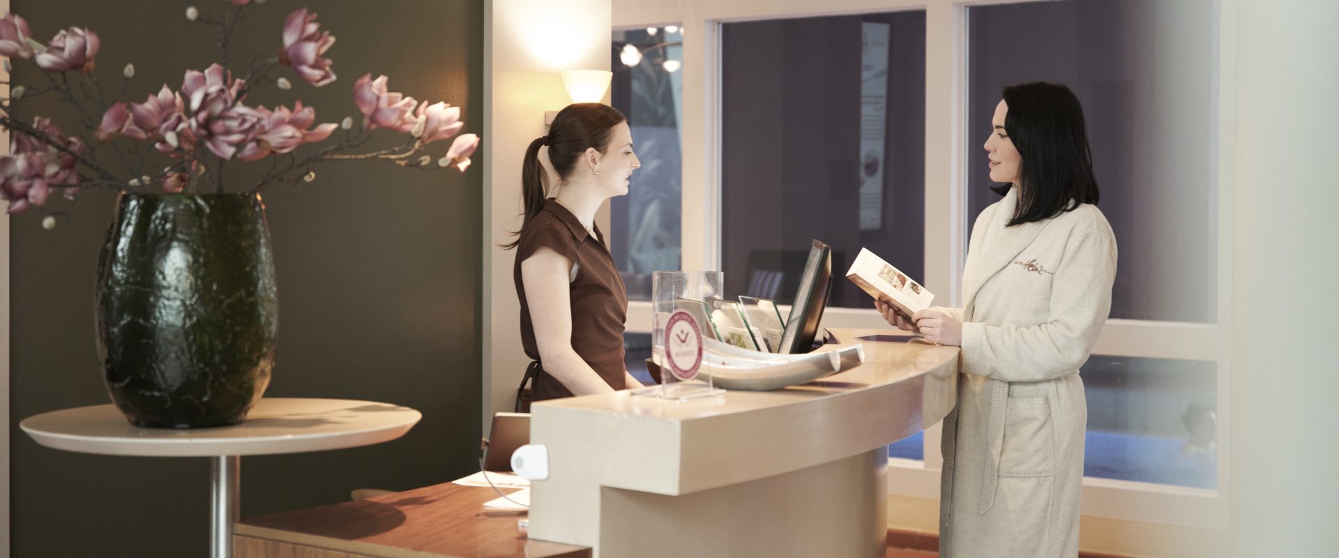 Zwei Frauen unterhalten sich an der Rezeption eines Spas. Eine im Bademantel hält eine Broschüre in der Hand, die Rezeptionistin lächelt. Auf dem Tresen steht eine Vase mit Blumen.
