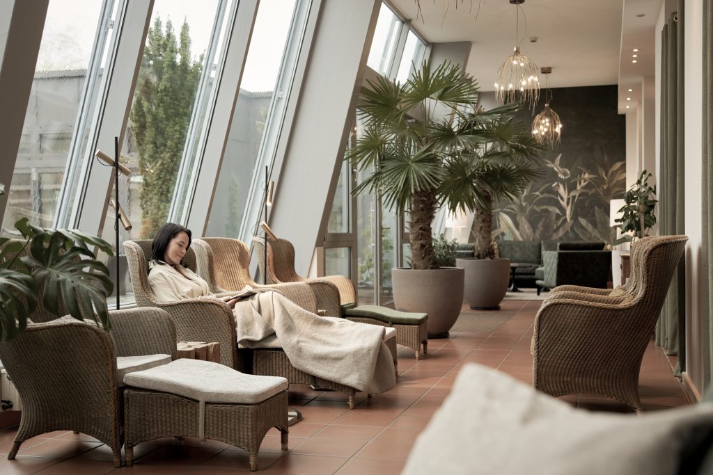 Eine Frau entspannt mit einer Decke in einer sonnendurchfluteten Lounge mit Korbstühlen, Pflanzen und großen schrägen Fenstern.