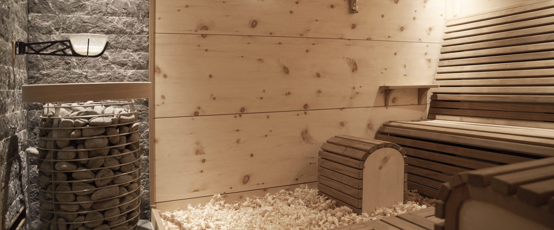 Moderne Holzsauna mit Bänken, Steinofen, Akzentsteinwand und Holzspänen auf dem Boden, sanft beleuchtet durch eine Wandlampe.